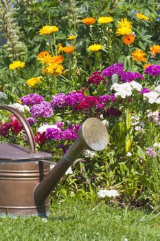 Old copper watering can stands in front of a colorful bed of summer flowers