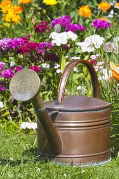 Old copper watering can stands in front of a colourful summer bed
