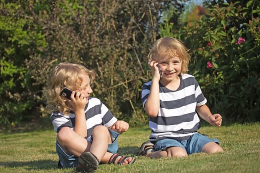 Two smiling blonde boys are sitting on the lawn and telephone.
