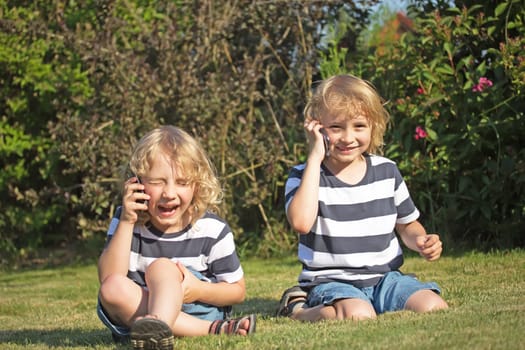 Two laughing  blonde boys are sitting on the lawn and telephone.