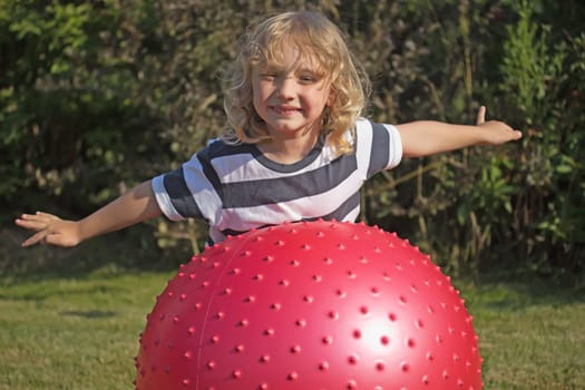 Attractive blond boy is playing with red gymnastic ball outdoors