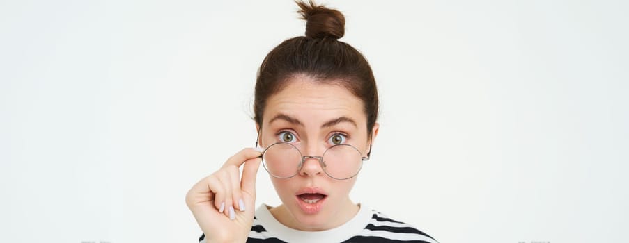 Image of woman in glasses, looks surprised, takes off eyewear, looks amazed, says wow, impressed by something, stands over white background.