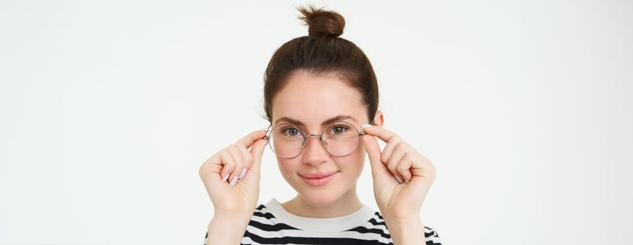 Close up portrait of smiling, attractive young woman wearing new eyewear, trying on glasses for vision, standing over white background.