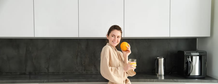 Portrait of young and healthy woman starts her day with fruits, eating fresh orange, drinking juice in kitchen, wearing bathrobe.