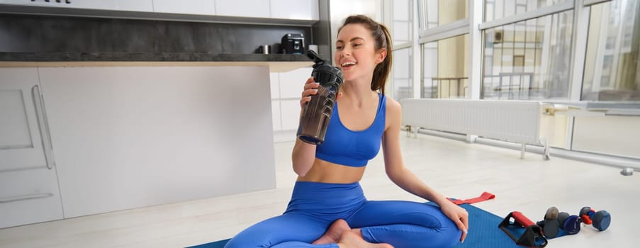 Young woman working out from home, drinking water during her sport training session in living room, sitting on yoga mat.