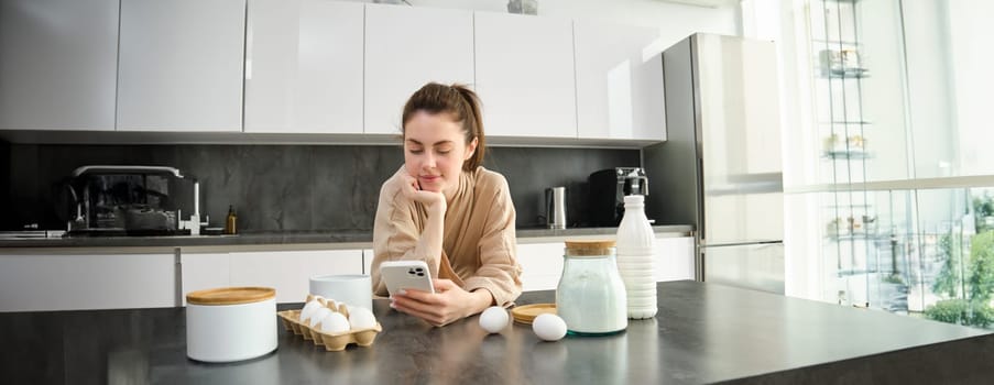 Attractive young cheerful girl baking at the kitchen, making dough, holding recipe book, having ideas.