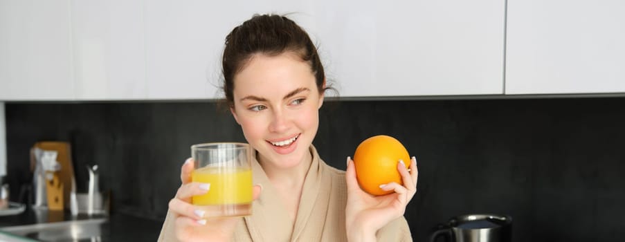 Image of good-looking healthy woman in bathrobe, drinking fresh juice, showing orange fruit, posing in kitchen.