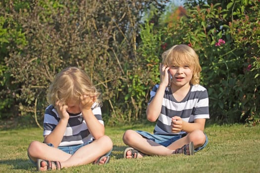 Two smiling blond boys are sitting on the lawn in the garden. One of them holding his head in his hands. The other is calling.