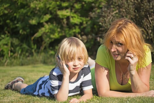 Mother with her blond hair son are lying and calling on the lawn in the garden.