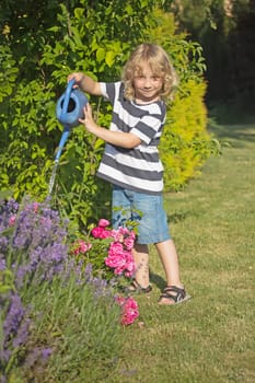 The blond boy is watering by blue pot flowers in the garden. Front view.