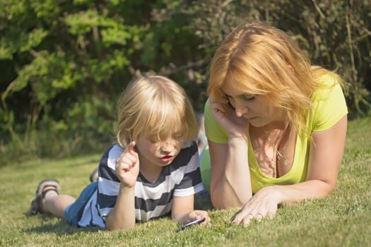 Mother with her blond hair son are lying in the garden. She is calling and her son is playing with smart phone.