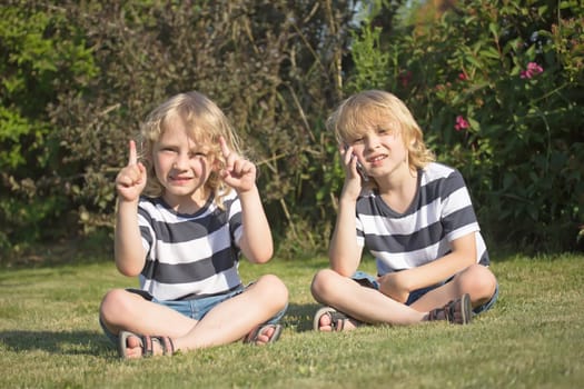 Two smiling blond boys are sitting on the lawn in the garden. One of them showed the fingers upward. The other is calling.