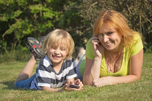 Laughing mother with her blond hair son are lying and calling on the lawn in the garden.