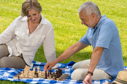 Retired senior couple playing chess in park sitting blanket grass