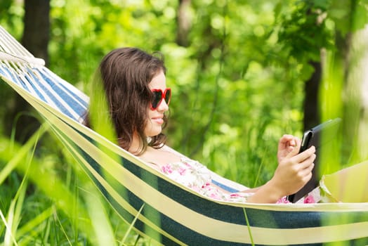 young woman lying in a hammock in the garden with e-book on tablet computer.
