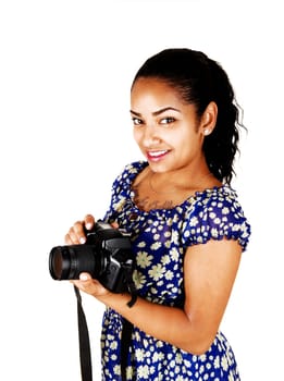 A young black woman in a blue dress standing for white background with.a camera in her hands, smiling..