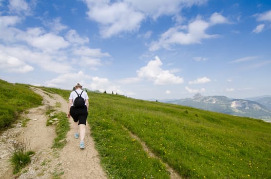 Female hiker on a path along a meadow with mountains and blue skies