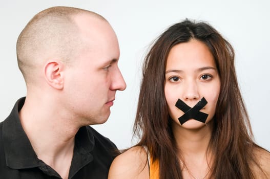 frontal head and shoulder shot of a young couple against a white background  woman with adhesive tape glued over crosswise mouth and sad look  Man in profile with an evil eye in the direction of woman