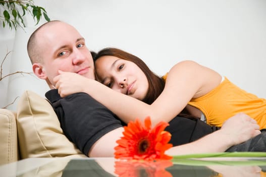 Upper body portrait of a young couple at home lying arm in arm on the couch with a single flower lying on the table in front of it