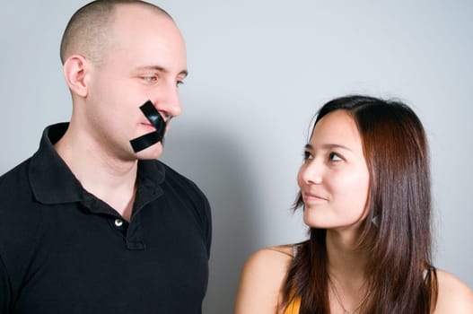 Head-and-shoulder photograph of a young couple in front of a bright wall  Man has over glued mouth  Woman looks smiling towards the man