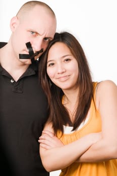 Upper body portrait of a young couple against a white background  Man has mouth glued crosswise with duct tape  Woman looks mischievously smiling with crossed arms into the camera