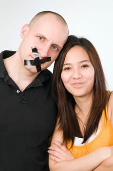 Upper body portrait of a young couple against a white background  Man has mouth glued crosswise with duct tape  Woman looks mischievously smiling with crossed arms into the camera
