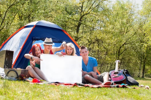 Four teenagers in colored shirts on the camping with a white board which can be used for your advertisements