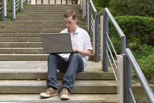 A young man with a laptop sits on a staircase and works.