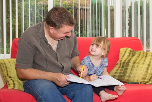 Father and daughter sitting on a sofa, drawing a picture together. The little girl is smiling up at her dad.
