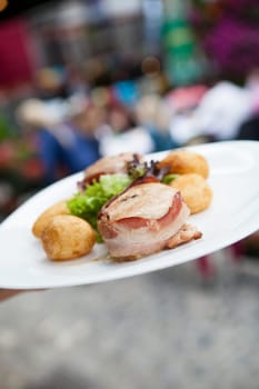 Closeup of grilled chicken breast wrapped in bacon with sides of baked potatoes and salad on white plate