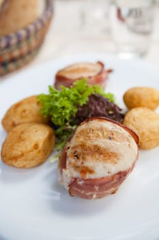 Closeup of grilled chicken breast wrapped in bacon with sides of baked potatoes and salad