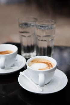 Two milk based coffees in white cups and glasses of water on cafe table