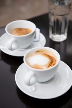 Closeup of two milk based coffees in white cups on cafe table with glass of water in background
