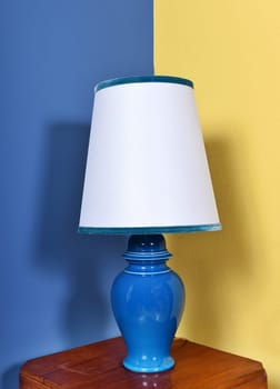 Blue ceramic lamp with simple white lampshade with matching blue trim standing on a table in the corner of a bi-color wall
