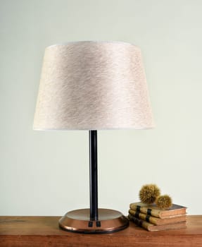 Classic lamp with white shade on black leg and shiny metal base, close-up against light wall, books and decorations on the shelf