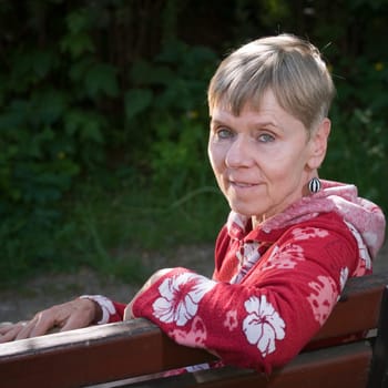 Portrait of an elderly woman on a park bench in summer with her upper body turned backwards over her backrest towards the camera