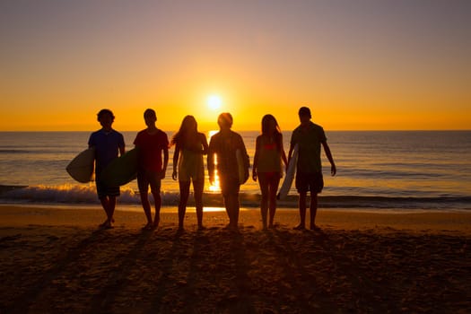 Surfers teen boys and girls group walking on beach at sunshine sunset backlight