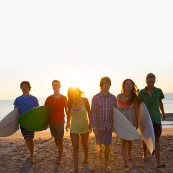 Surfers teen boys and girls group walking on beach at sunshine sunset backlight