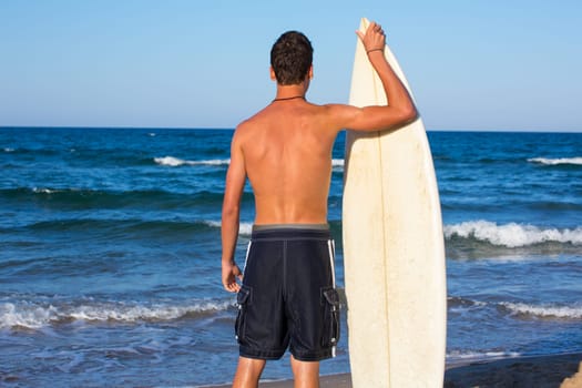 Boy surfer back rear view holding surfboard on blue beach