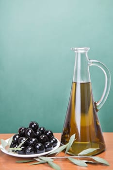 A jar with olive oil ans a plate with black olives on a wooden surface.