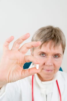 Head-and-shoulder shot of an elderly woman in a white coat holds a white medicine capsule between two fingers and looks into the camera