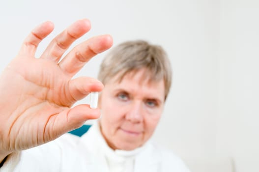 Head-and-shoulder shot of an elderly woman in a white coat holds a white medicine capsule between two fingers and looks into the camera