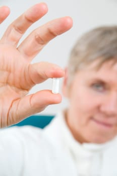 Head-and-shoulder shot of an elderly woman in a white coat holds a white medicine capsule between two fingers and looks into the camera