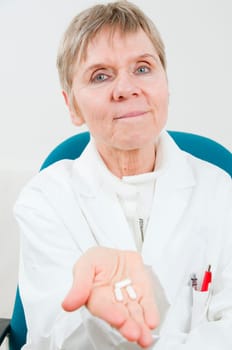 an elderly woman in a white doctor's coat shows three medicine capsules lying on her hand sitting on a chair