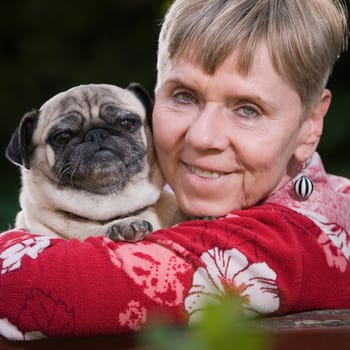 Head-and-shoulders portrait of a mature woman in the park with a dog (pug) in her arms and a smiling look into the camera