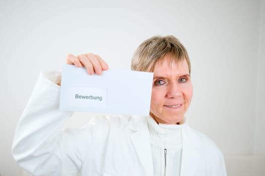 Upper body shot of an elderly woman in a white doctor's coat holds up an envelope with application inscription next to her