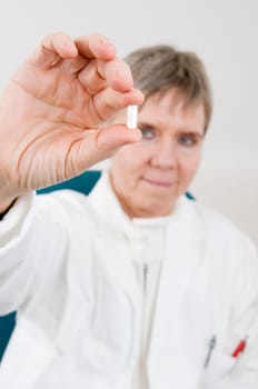 Head-and-shoulder shot of an elderly woman in a white coat holds a white medicine capsule between two fingers and looks into the camera
