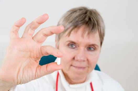 Head-and-shoulder shot of an elderly woman in a white coat holds a white medicine capsule between two fingers and looks into the camera