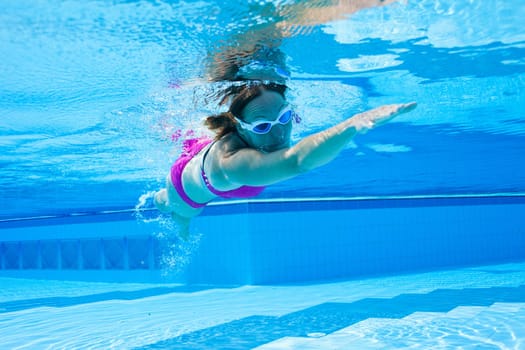 Underwater view of female with violet swimsuit and white goggles swimming in pool