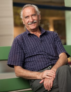 An older man (senior) sits relaxed on a bench,looking into the camera